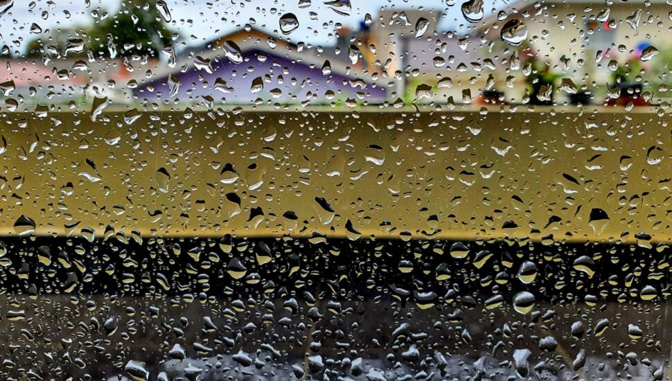 Imagem mostra janela com pingos da chuva e casas ao fundo