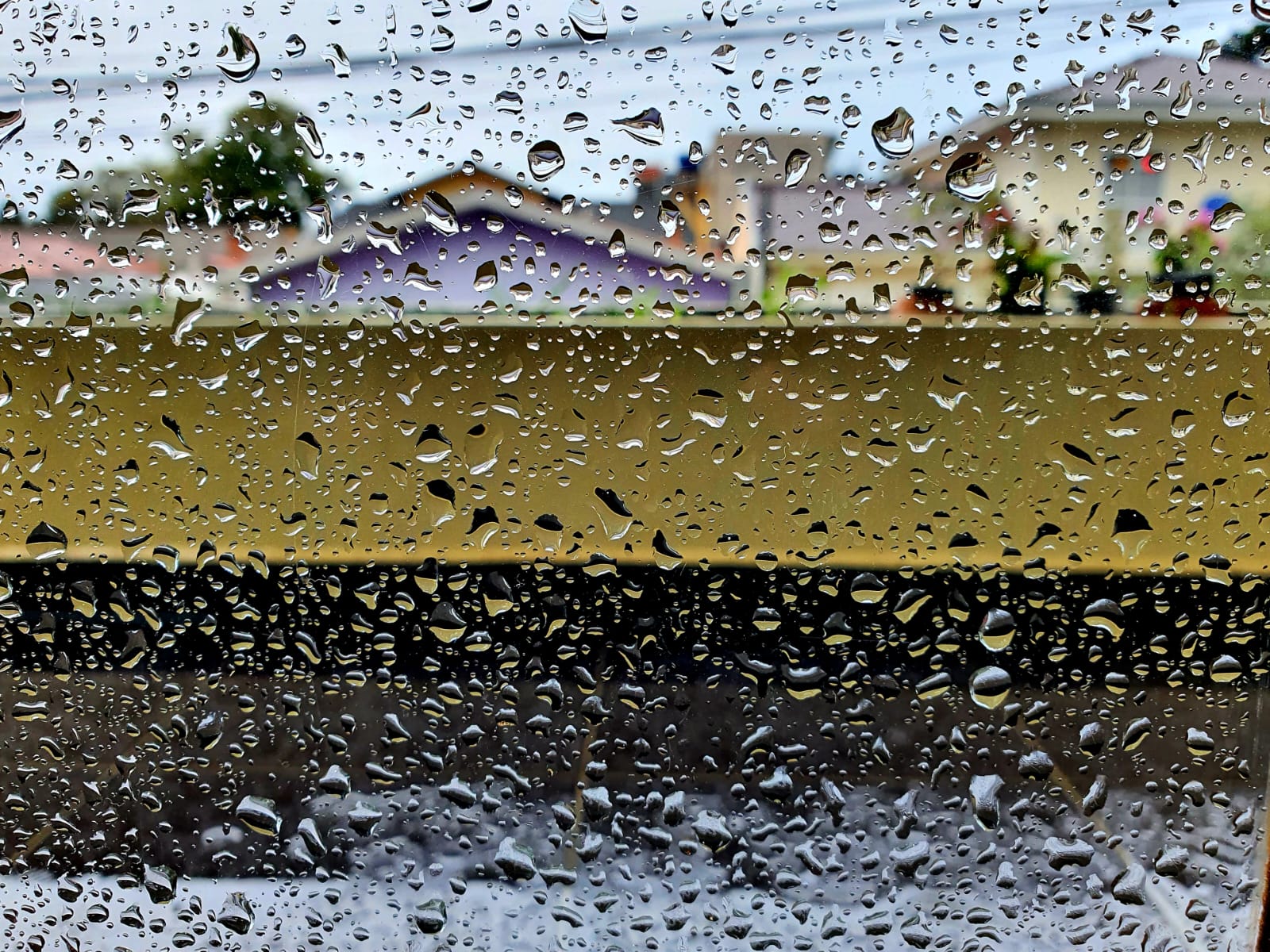 Imagem mostra janela com pingos da chuva e casas ao fundo