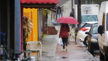 Imagem mostra a chuva que cai em Curitiba. uma mulher aparece com um guarda chuva e caminhando em calçadas com varias poças de água,