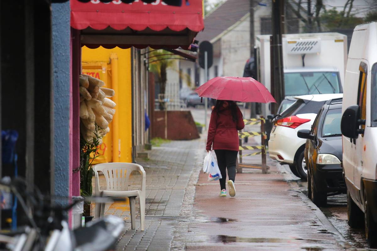 Imagem mostra a chuva que cai em Curitiba. uma mulher aparece com um guarda chuva e caminhando em calçadas com varias poças de água,