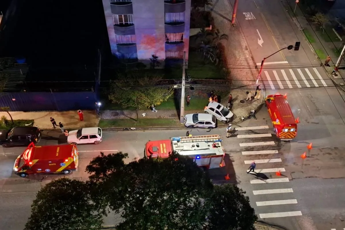 Imagem de cima mostra um acidente de trânsito com dois carros brancos e viaturas dos bombeiros.