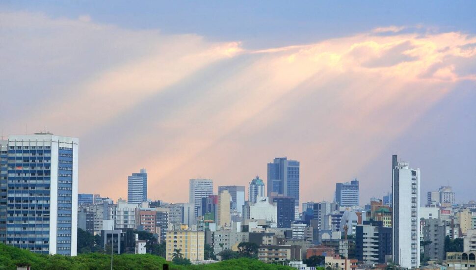 Imagem mostra Curitiba com céu fechado e os raios de sol passando pelas nuvems.