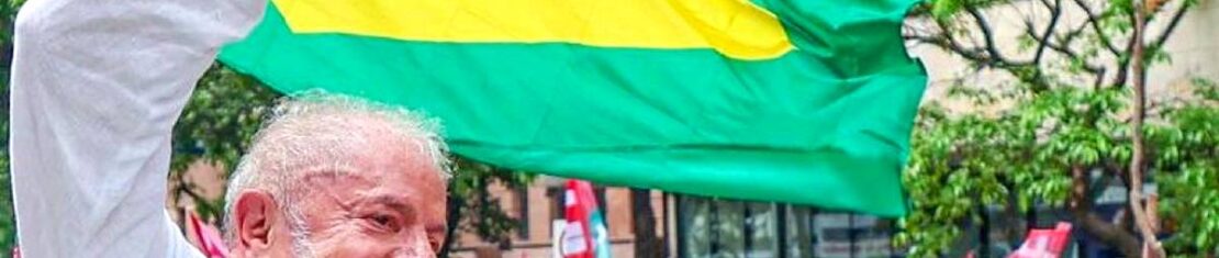 Imagem mostra candidato Lula com multidão ao fundo e bandeira do Brasil em segundo plano