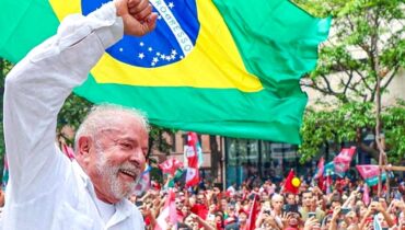 Imagem mostra candidato Lula com multidão ao fundo e bandeira do Brasil em segundo plano