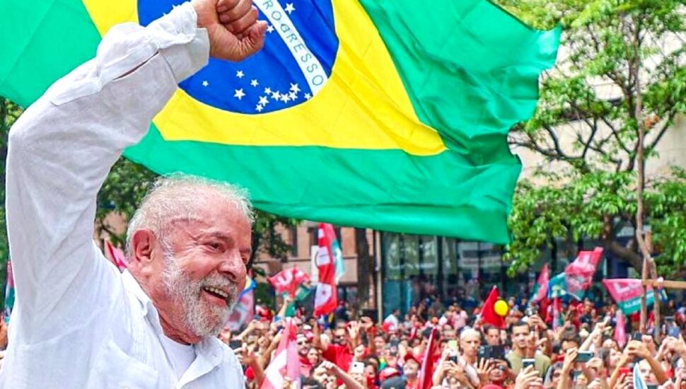 Imagem mostra candidato Lula com multidão ao fundo e bandeira do Brasil em segundo plano
