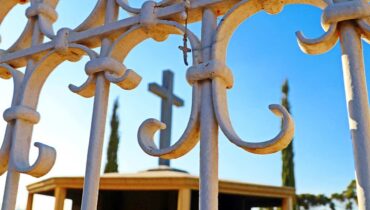 Cemitério em Curitiba: Cruz tem destaque atrás de uma grade num céu azul.