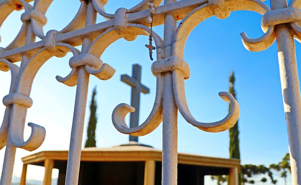 Cemitério em Curitiba: Cruz tem destaque atrás de uma grade num céu azul.