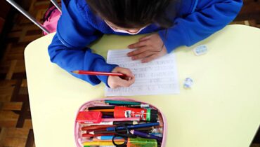 Imagem mostra uma criança escrevendo em um papel. A imagem é de cima e a criança esta com um estojo com material escolar e de uniforme azul.