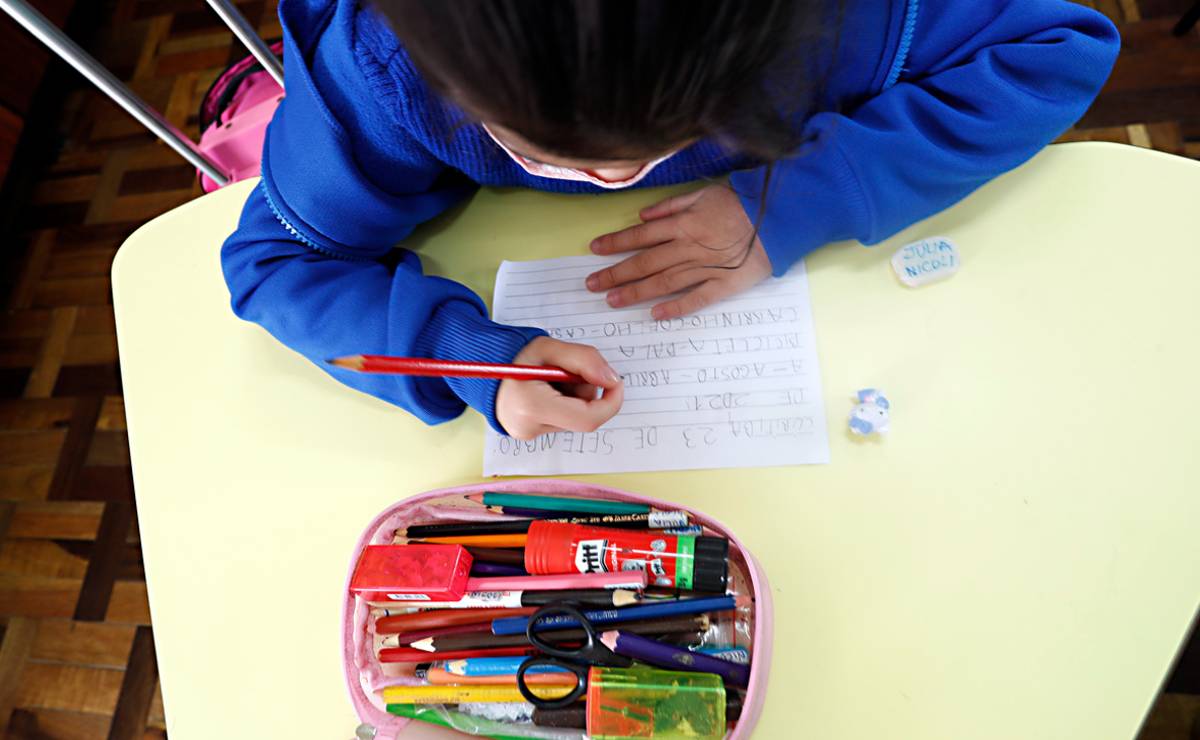 Imagem mostra uma criança escrevendo em um papel. A imagem é de cima e a criança esta com um estojo com material escolar e de uniforme azul.
