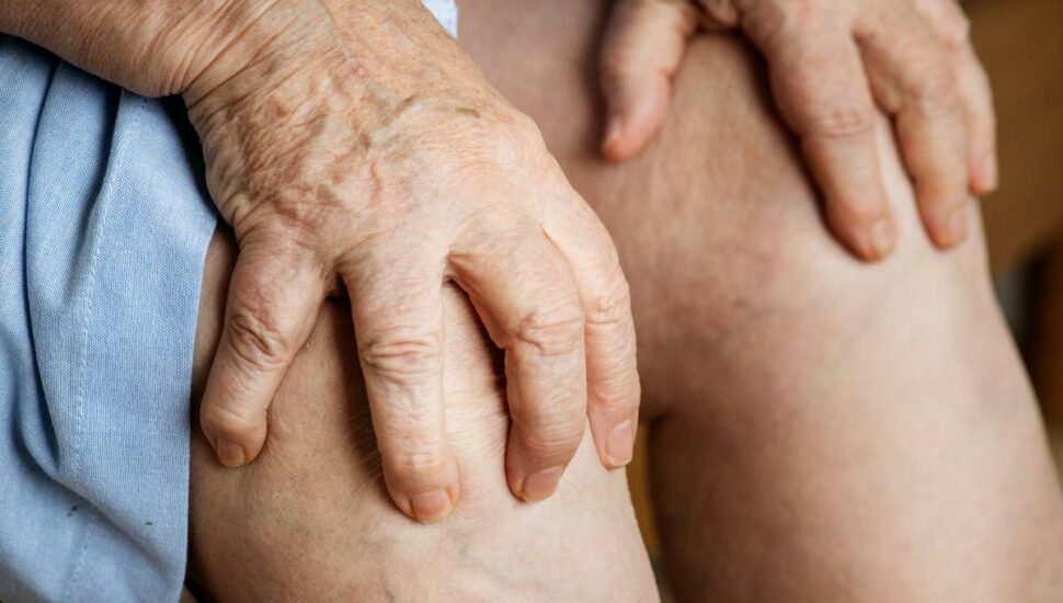 Imagem mostra uma idosa com as mãos no joelho, indicando dor na articulação