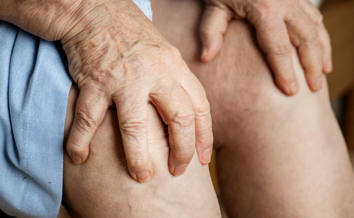 Imagem mostra uma idosa com as mãos no joelho, indicando dor na articulação