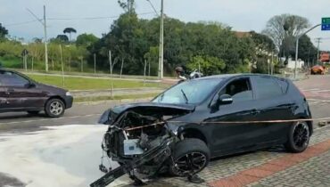 Imagem mostra um carro preto com a frente destruída e espuma anti-incêndio no chão