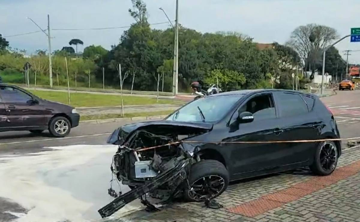 Imagem mostra um carro preto com a frente destruída e espuma anti-incêndio no chão