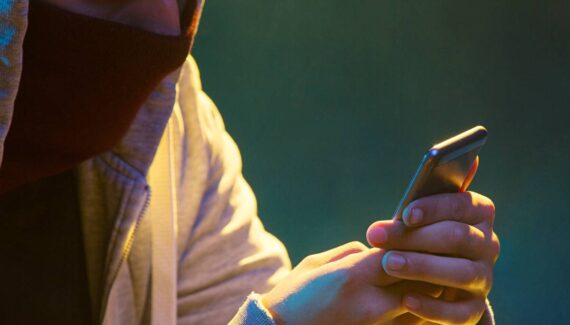 Imagem mostra um homem com uma máscara mexendo em um celular numa sala escura