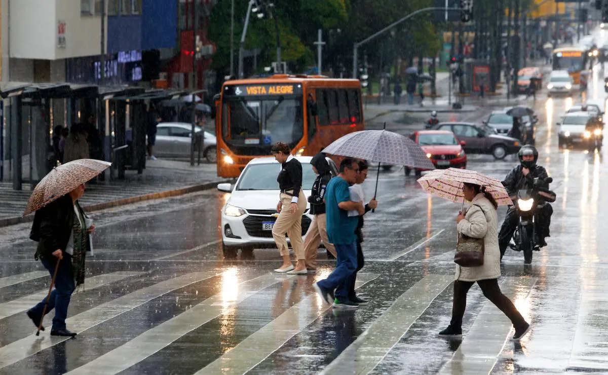 Imagem mostra pessoas ultrapassando uma grande avenida na faixa de pedestres e muita chuva caindo. Todos estão de guarda chuva.