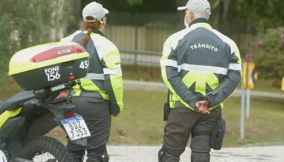 Imagem mostra dois agentes de trânsito de Curitiba de costas orientando o transito com uma moto ao lado estacionada