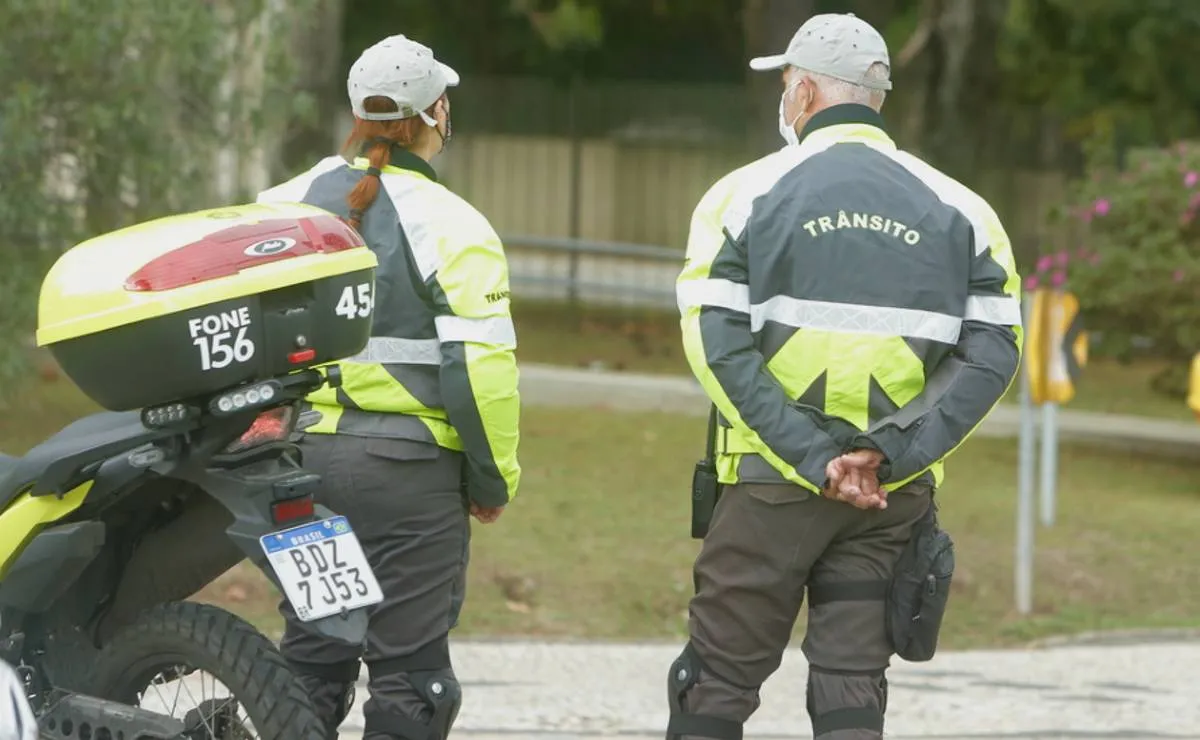 Imagem mostra dois agentes de trânsito de Curitiba de costas orientando o transito com uma moto ao lado estacionada