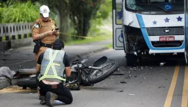 Imagem mostra um acidente de trânsito em Curitiba, com uma moto caída no asfalto e dois policiais fazendo medições. Ao fundo um ônibus com o para choque quebrado
