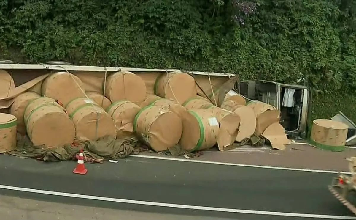 Imagem mostra um caminhão tombado com bobinas de papel espalhadas pela pista.