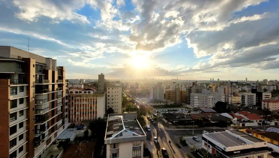 Imagem mostra o pôr do sol em Curitiba, com o sol entre nuvens e prédios.