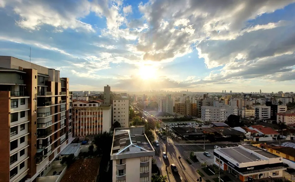 Imagem mostra o pôr do sol em Curitiba, com o sol entre nuvens e prédios.