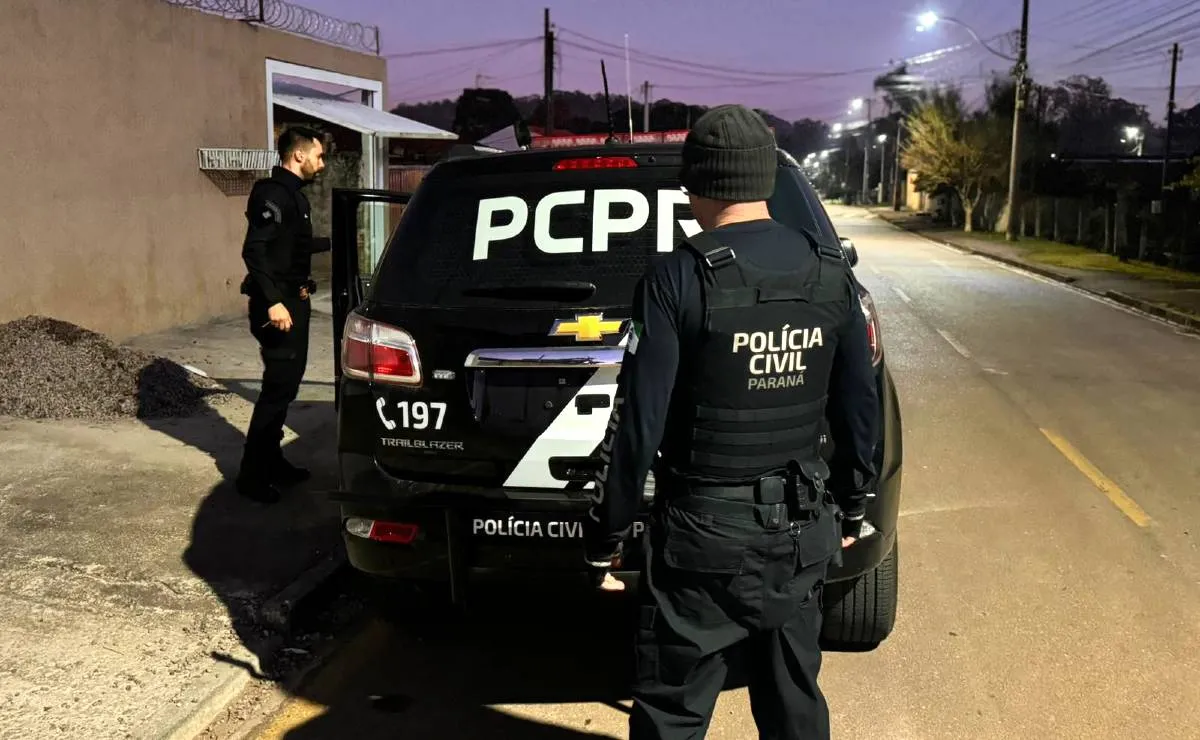 Imagem mostra uma viatura da Polícia Civil do Paraná durante a operação em uma rua, a noite, em Piraquara. Dois policiais estão de costas.