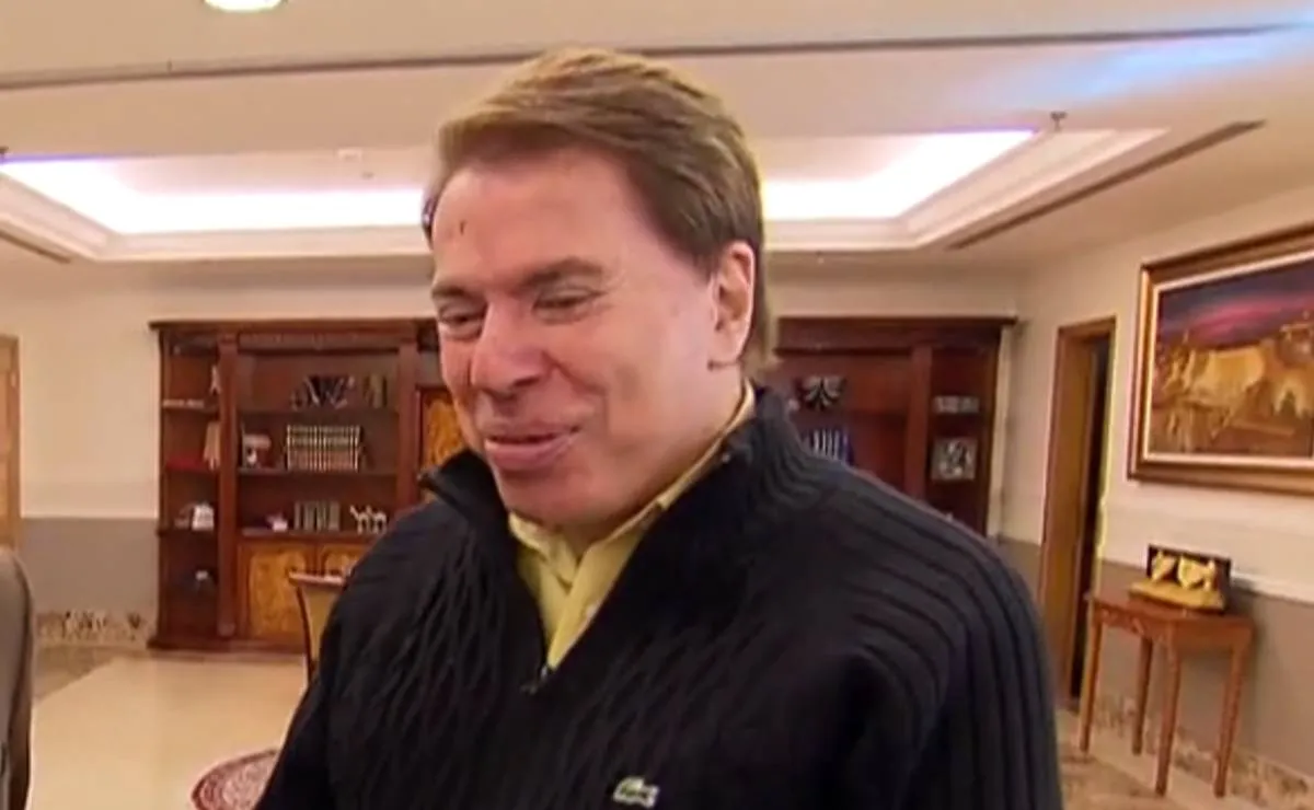 Imagem mostra Silvio Santos em uma sala com uma blusa de la preta e uma camisa cor de mostarda.
