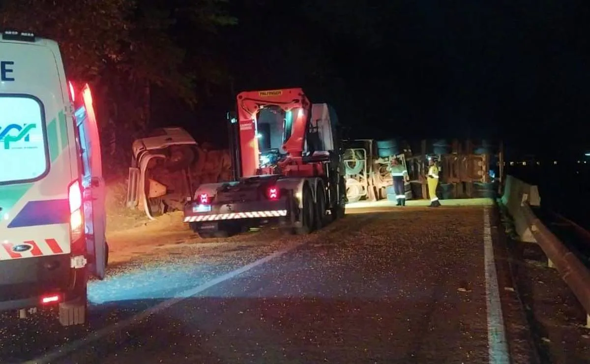 Imagem a noite mostra um caminhão tombado em uma rodovia, uma ambulância do lado esquerdo e luzes de farol.