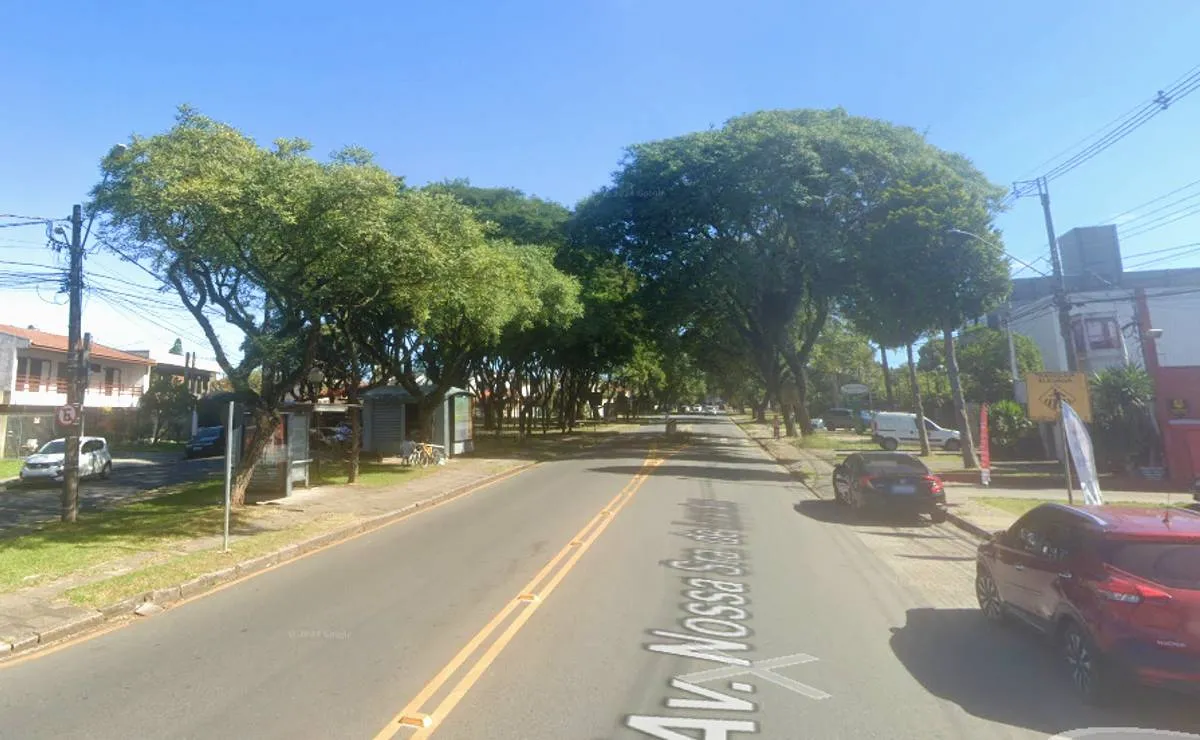 Imagem mostra uma avenida de Curitiba com asfalto e árvores grande. Tem um carro vermelho estacionado no lado direito da imagem.