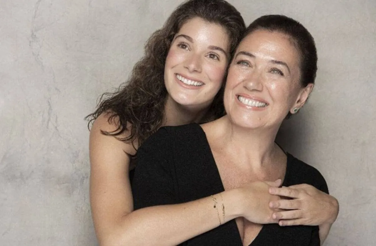 imagem mostra a atriz lilia cabral em frente a um fundo conza, de orupa preta, sendo abraçada pela filha giulia