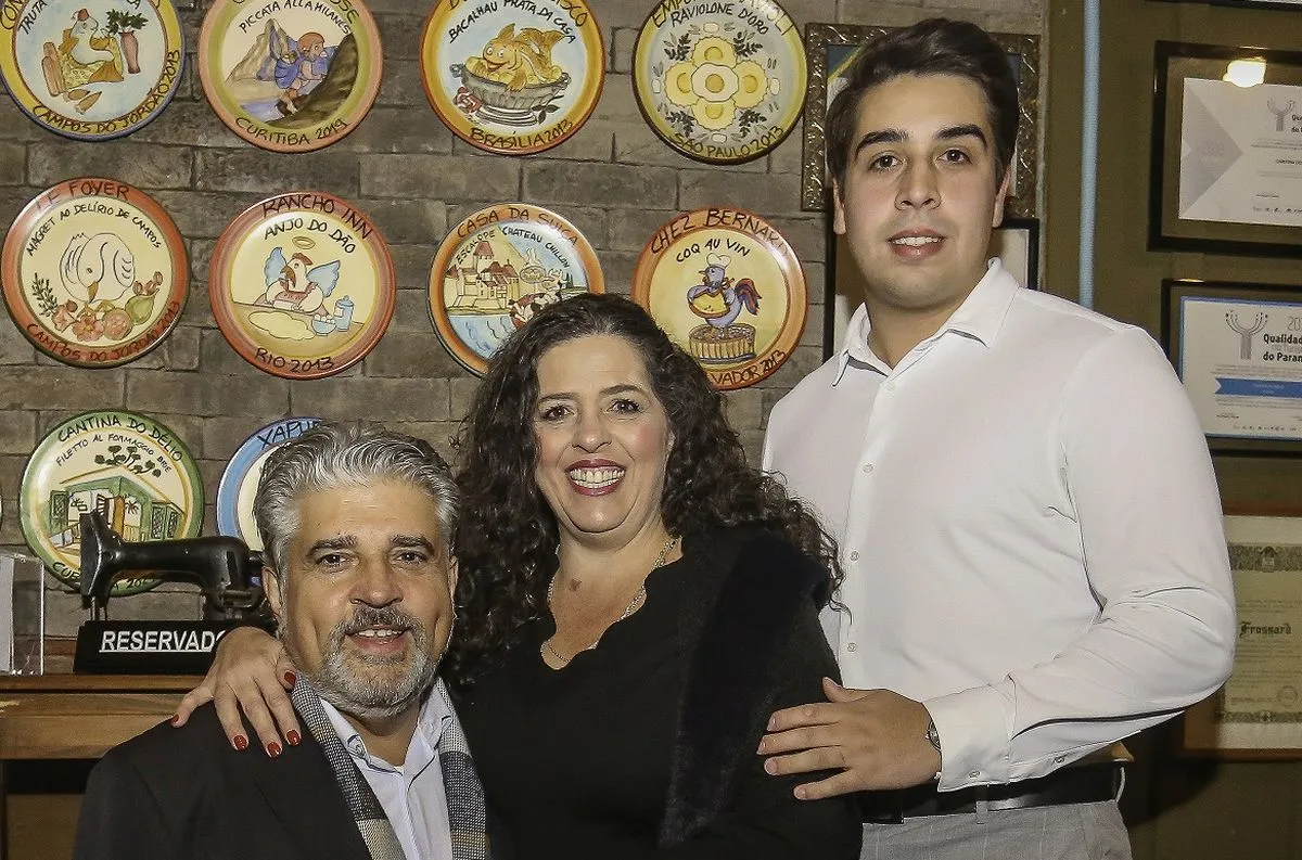 Uma parede com vários pratos pendurados e a na frente os empresários Délio, Renata e César abraçados