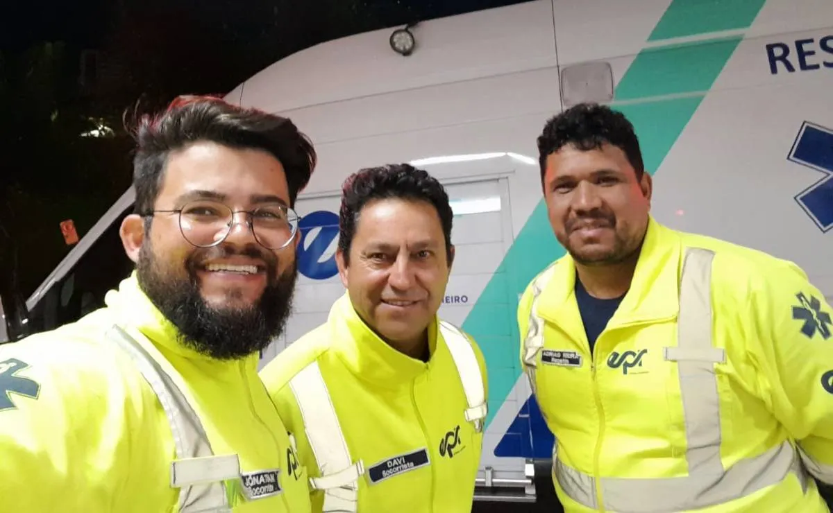 Imagem mostra três socorristas em frente a uma ambulância.