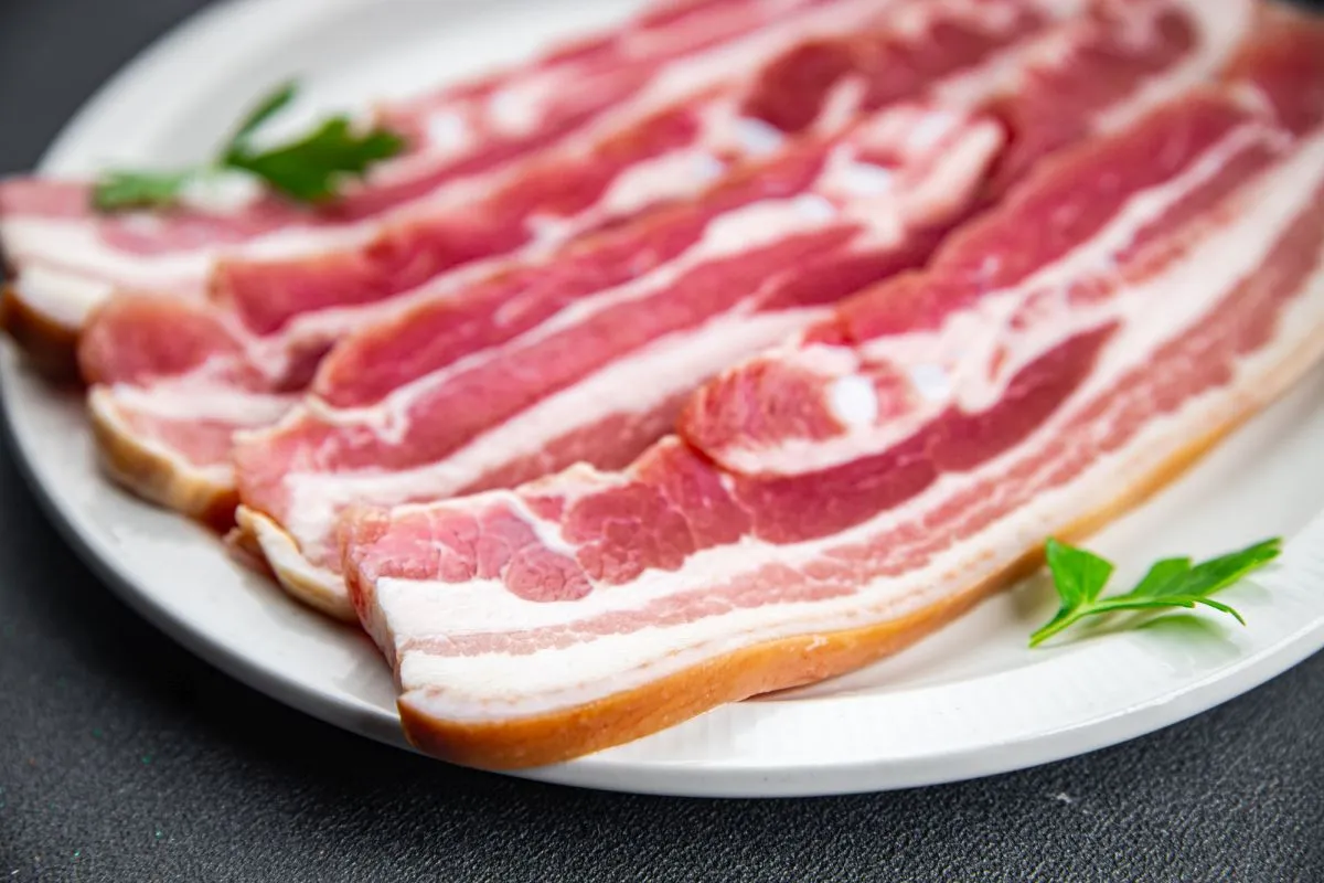 imagem mostra quatro fatias de bacon cru em um prato decorado com ervas