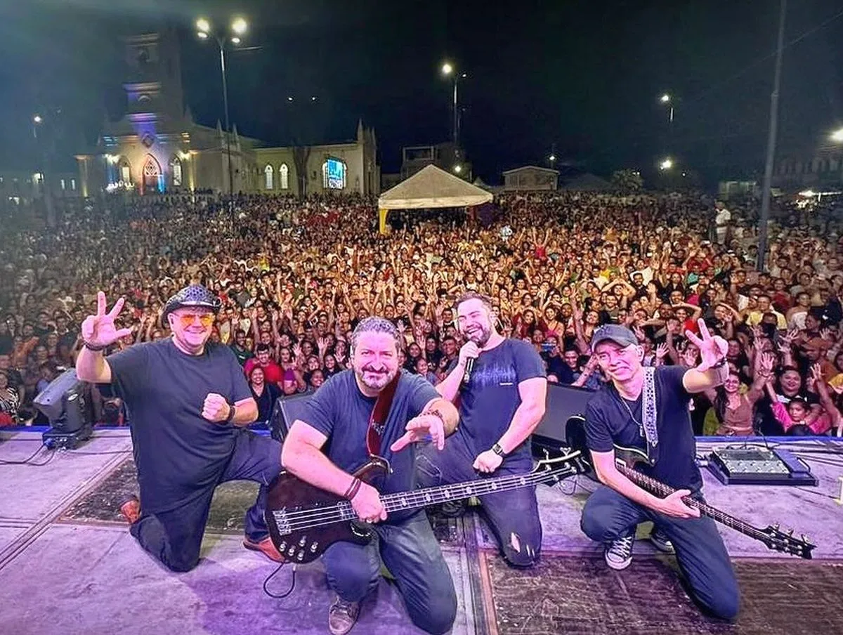 imagem mostra integrantes da banda no palco de um show, com o público ao fundo