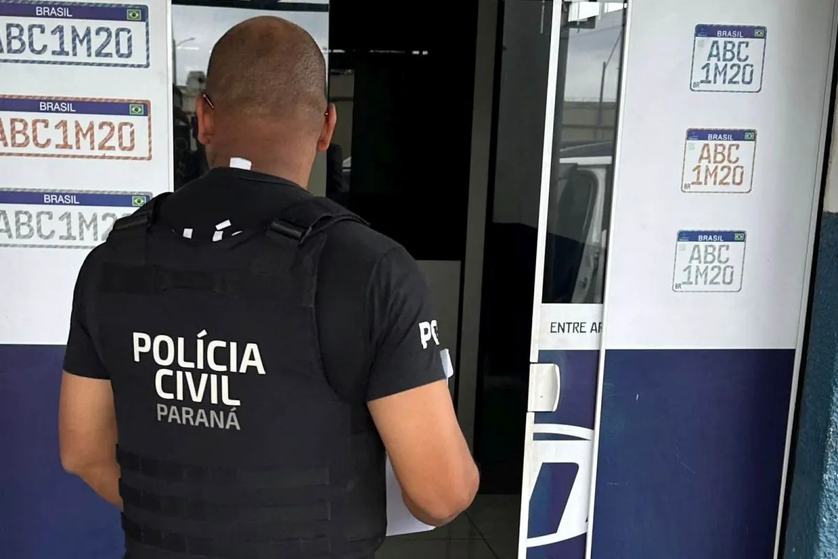 Na imagem, policial em frente à porta de entrada de uma empresa de emplacamento de veículos.