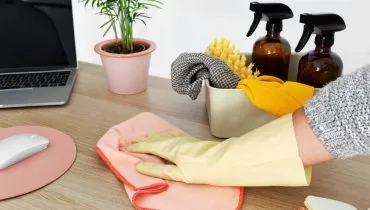 Imagem mostra uma pessoa passando um pano de limpeza em uma mesa com um computador e dois borrifadores.