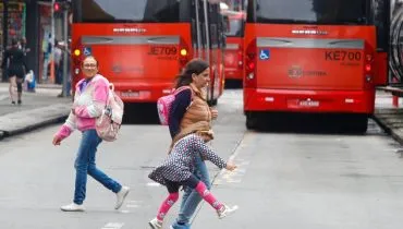 Imagem mostra dois biarticulados em Curitiba com pedestres atravessando a rua. Um dos pedestres é uma criança travessa.