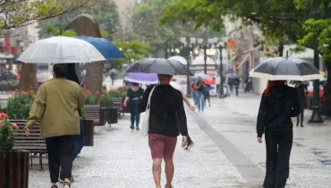 Na imagem, pessoas caminham na chuva, com guarda-chuvas, no calçadão da Rua XV.