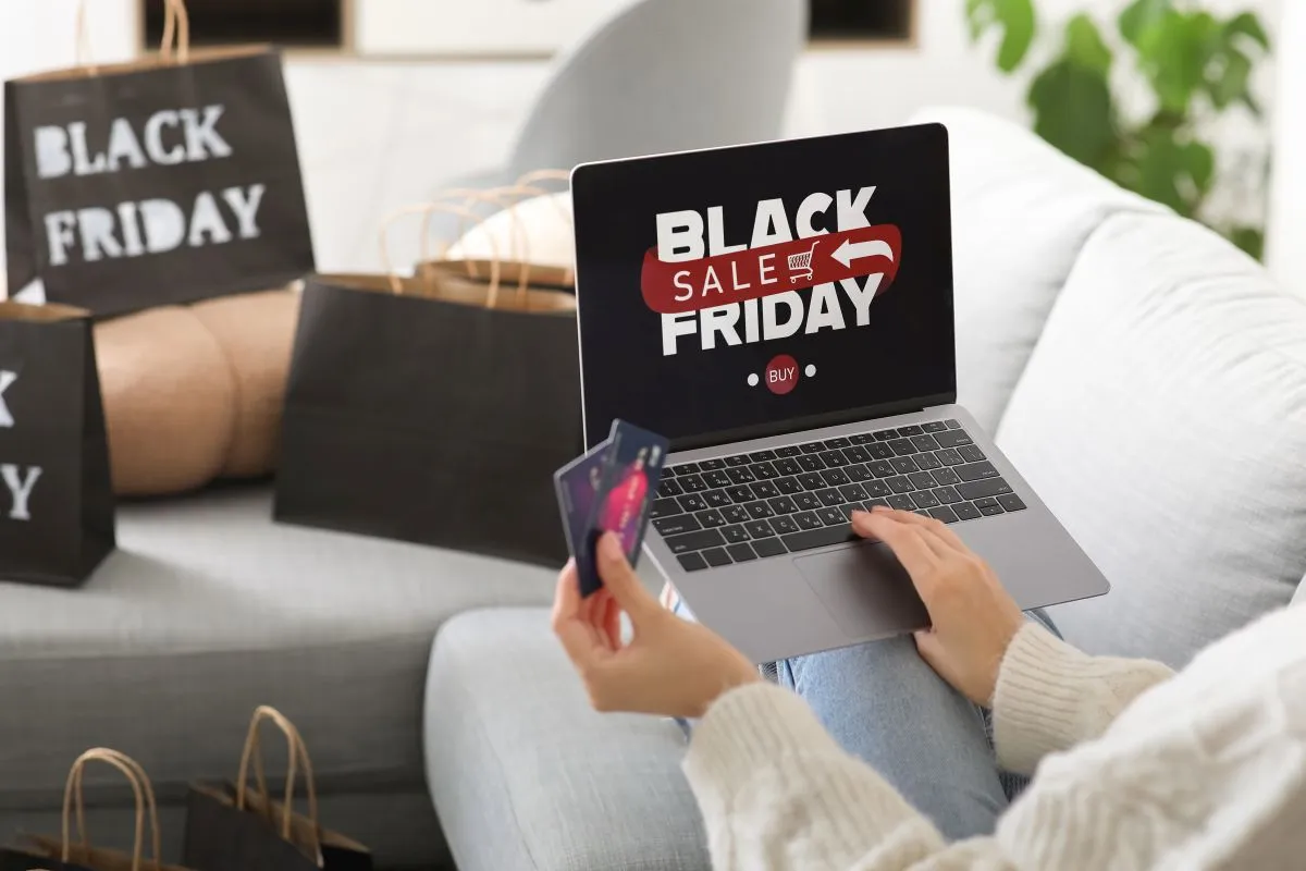 Na imagem, uma pessoa segura um laptop com "Black Friday" na tela. Há sacolas de compras sobre um sofá no ambiente.