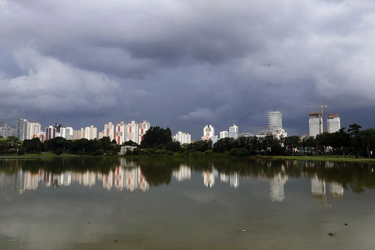 Na imagem, nuvens de chuva sobre prédios em Curitiba e lago de parque