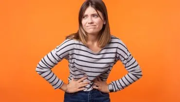 Imagem mostra uma mulher com dor de barriga, Ela está em um fundo laranja usando blusa listrada e calça preta.