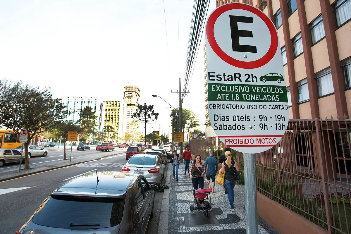 imagem mostra placa com explicações sobre o Estar em primeiro plano e carros estacionados ao longo de um via de Curitiba. Pessoas passeiam pela calçada