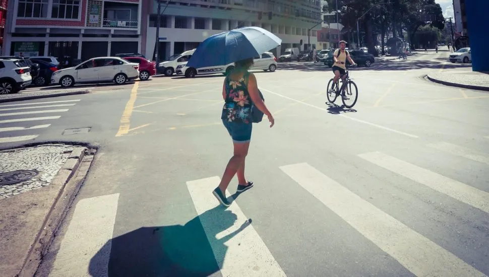 Imagem mostra pessoas andando na rua e mulher usando sombrinha para se proteger do calor