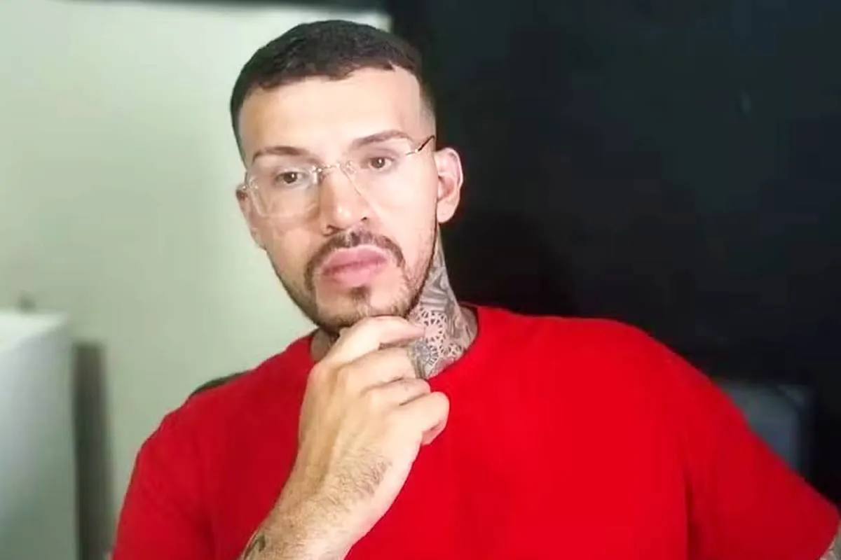 Imagem mostra um homem com uma blusa vermelha, óculos, tatuagem e barba por fazer.