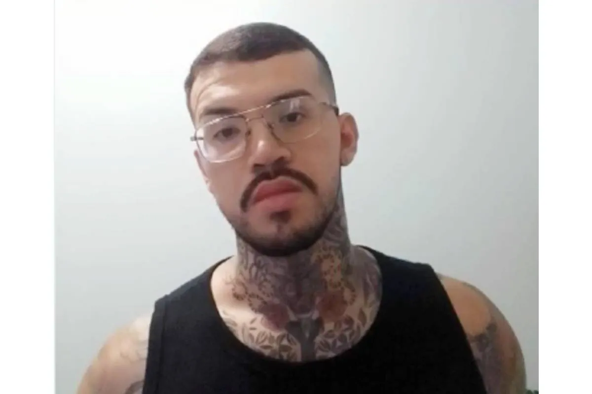 Imagem mostra um homem com uma camisa preta, óculos, tatuagem e barba por fazer.