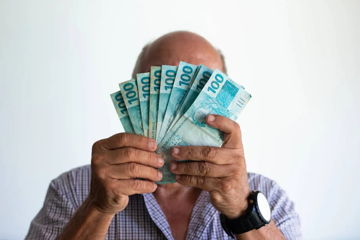 Imagem mostra um homem escondendo o rosto atrás de um leque com notas de R$ 100