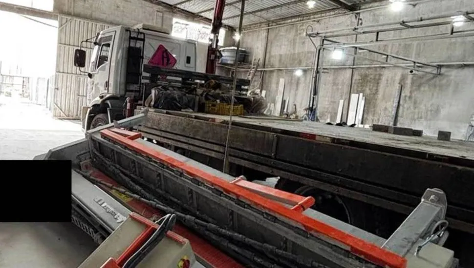 Imagem mostra um caminhão com uma máquina pesada caída ao lado, dentro de um barracão.