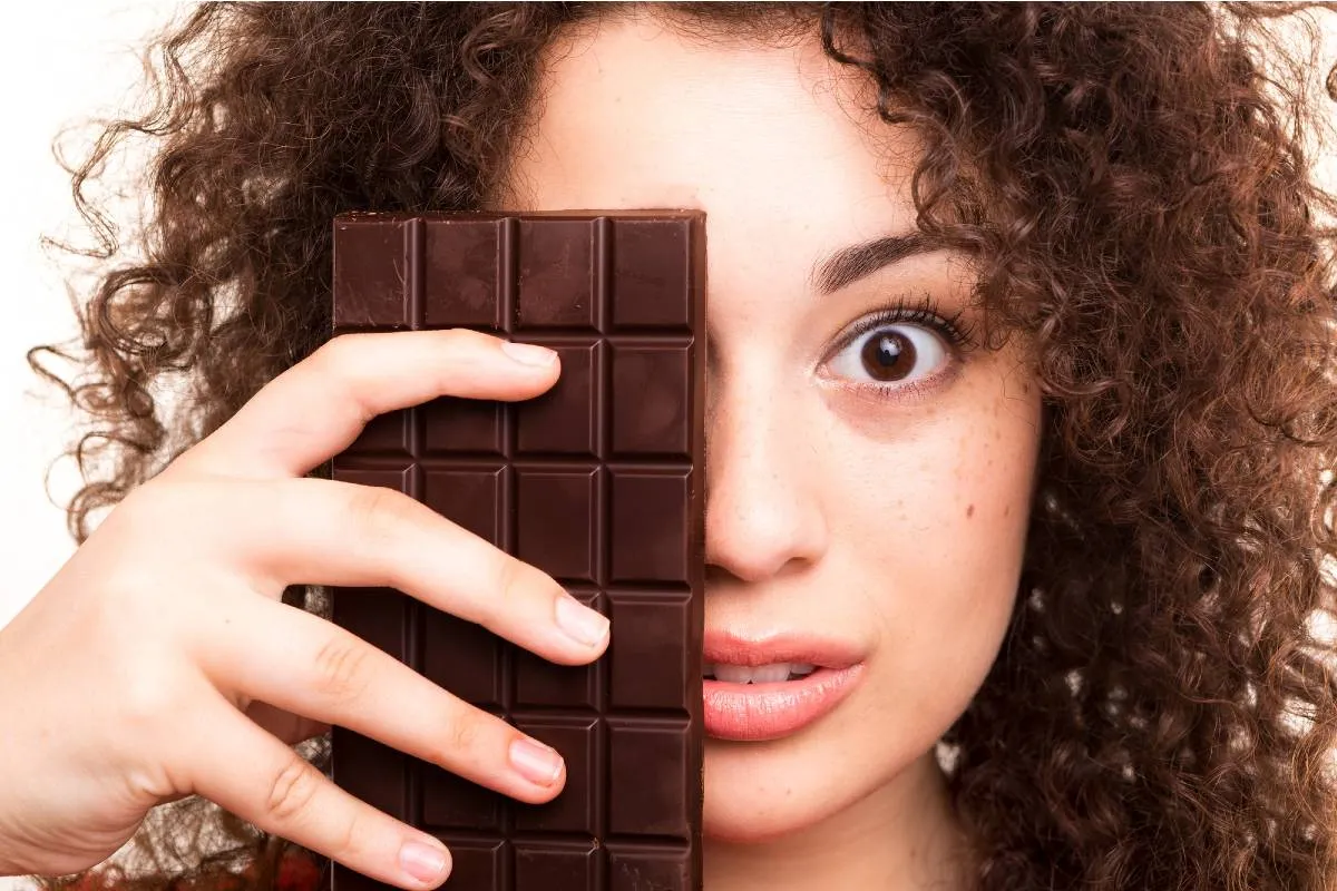 Imagem mostra uma mulher com cabelos cacheados tampando o rosto com uma barra de chocolate.