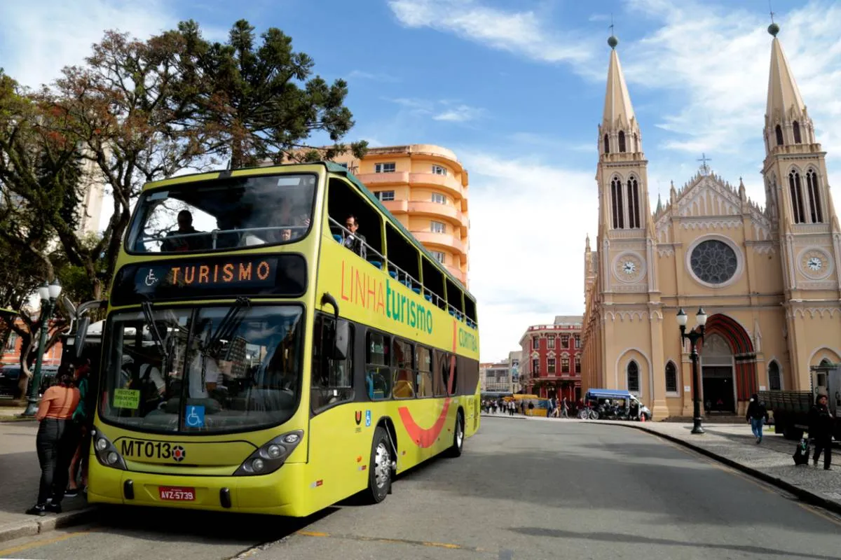 Imagem mostra um ônibus verde, de dois andares, em frente à catedral de Curitiba
