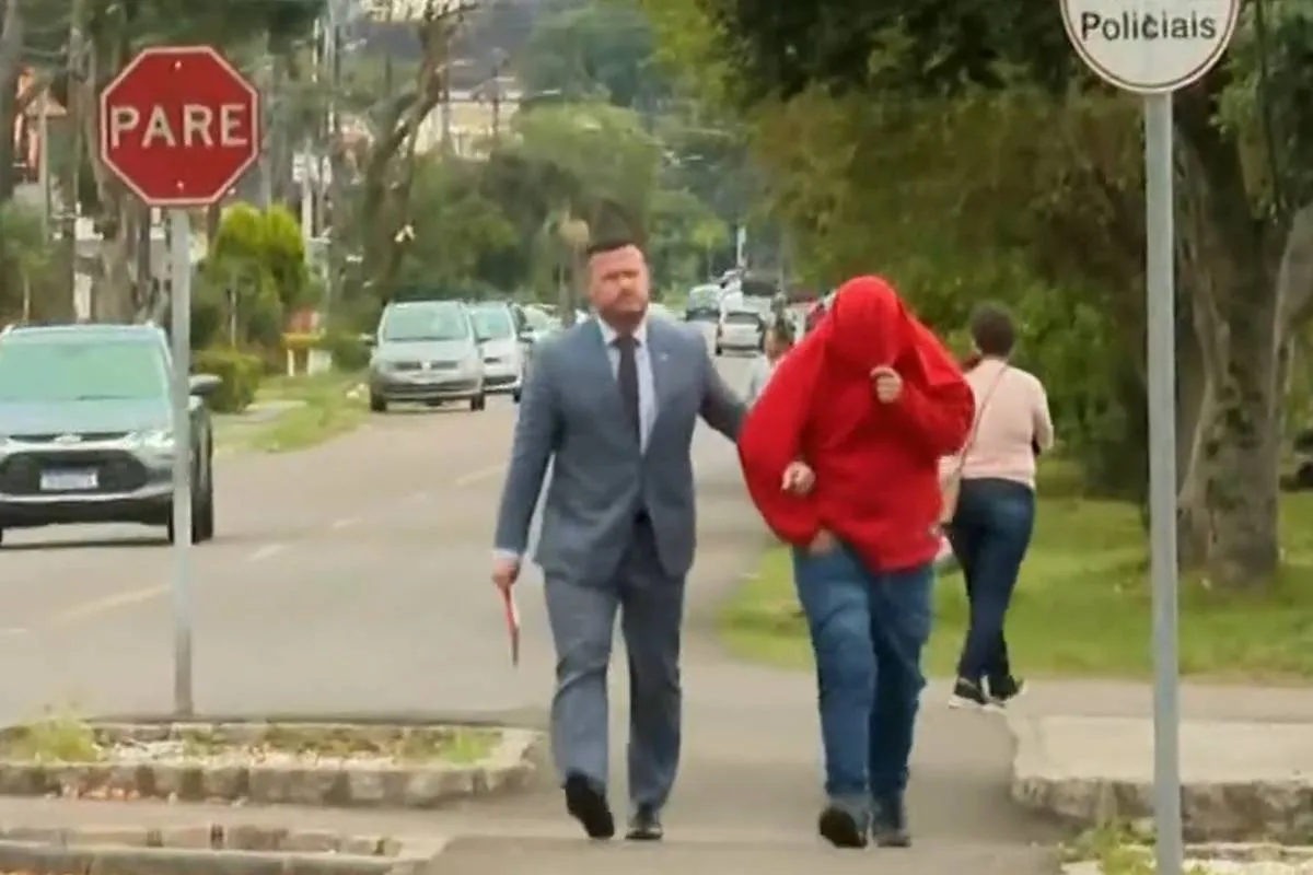 Imagem mostra um homem de terno guiando um homem de blusa vermelha e rosto coberto.